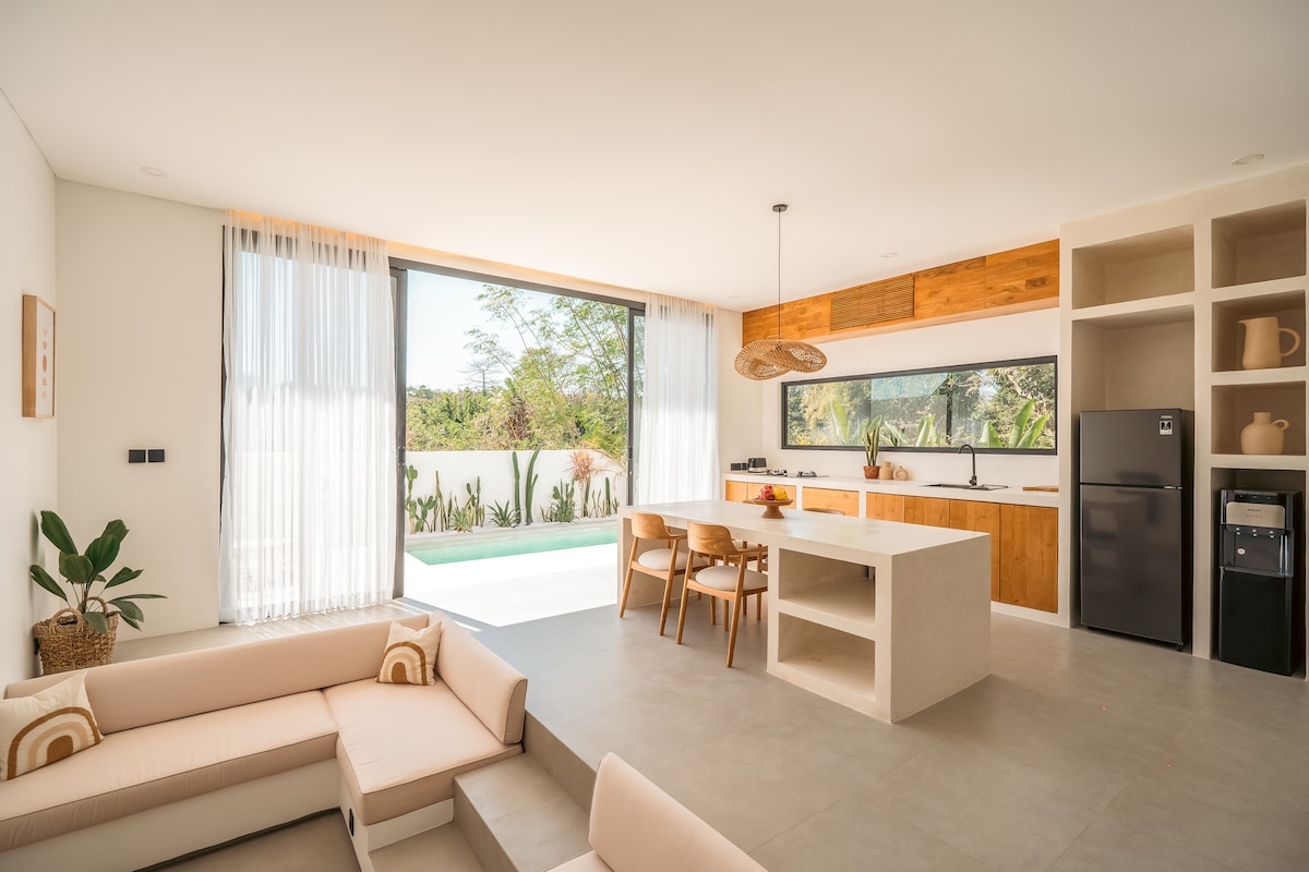 A modern living space features a light-colored sofa arranged around a flat surface table. Natural light floods in through large glass doors that open to the private pool area. A sleek kitchenette with wooden accents and essential appliances is visible in the background.
