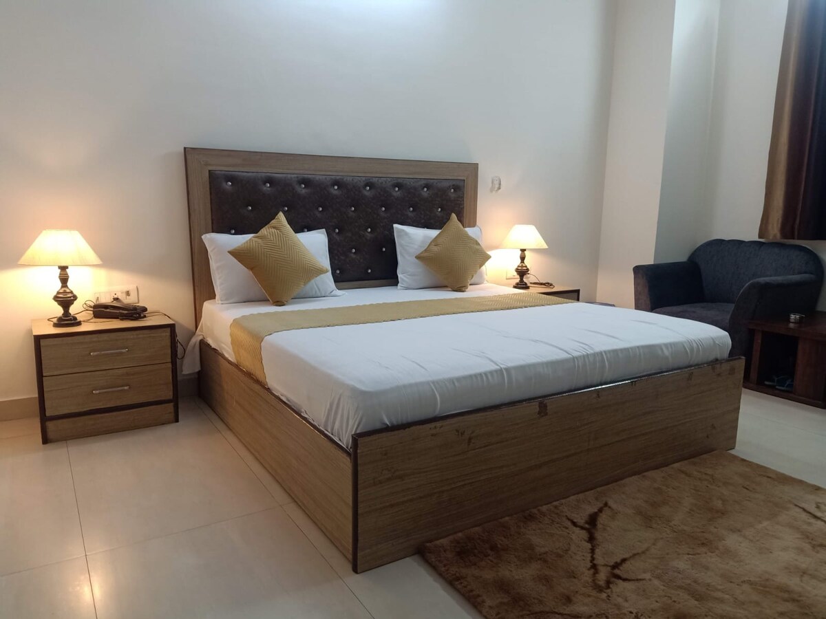 A spacious sleeping area is furnished with a wooden bed featuring a tufted headboard and decorative throw pillows. Two bedside lamps provide soft lighting, complementing a cozy armchair in the corner. A neutral-toned rug enhances the room's comfort while soft natural light enters through a nearby window.