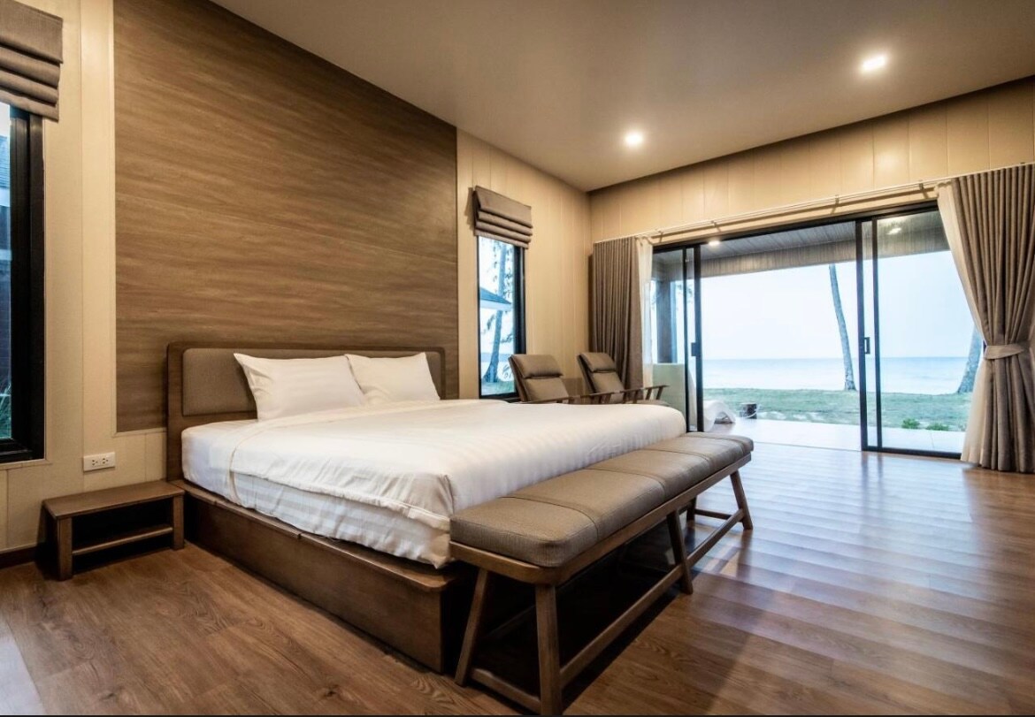 A spacious sleeping area features a large bed with a neutral-toned coverlet. Two benches are placed at the foot of the bed. Floor-to-ceiling glass doors open to a view of the outside, complemented by soft curtains and wooden flooring.