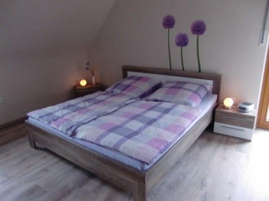 A spacious bed with a plaid duvet cover is flanked by two bedside tables, each holding a softly glowing lamp. The walls feature floral decals, and natural light enters through a window, creating a calm and inviting space.