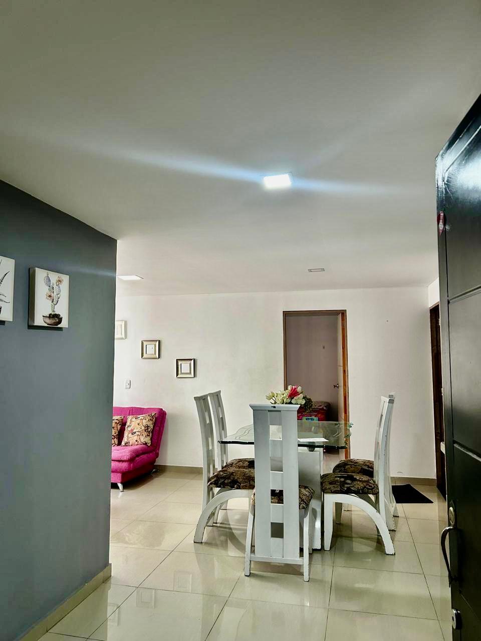 A modern dining area is visible, featuring a glass-top table surrounded by four chairs. Adjacent to the dining space, a cozy living area is suggested by a pink sofa adorned with decorative cushions. Light-colored walls enhance the sense of openness.