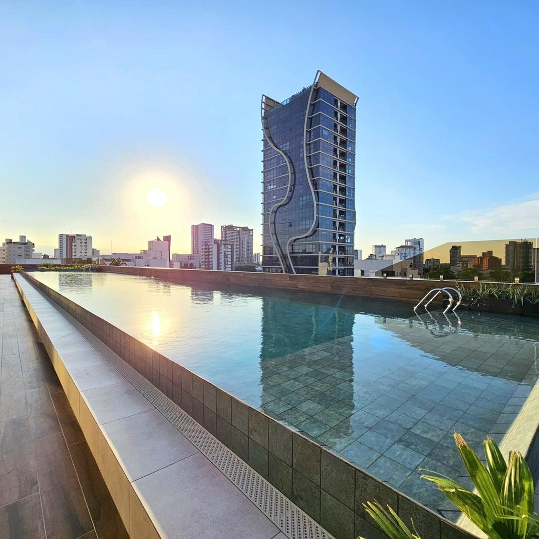 An infinity pool is shown reflecting the clear sky and sun, with a modern high-rise building in the background. Surrounding greenery adds a touch of nature to the sleek urban environment. The pool area is designed for relaxation and enjoyment.