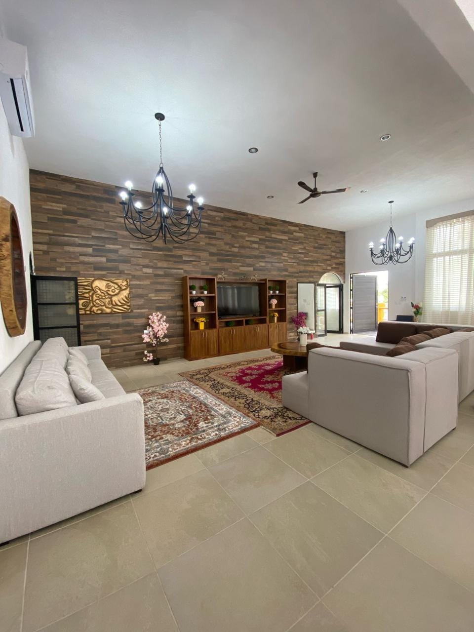 A spacious living area features a mix of light and dark furnishings, centered around a wooden entertainment unit. Two sofas and a reclined armchair are arranged on a soft area rug, with elegant chandeliers hanging from the ceiling. Natural light filters through large windows.
