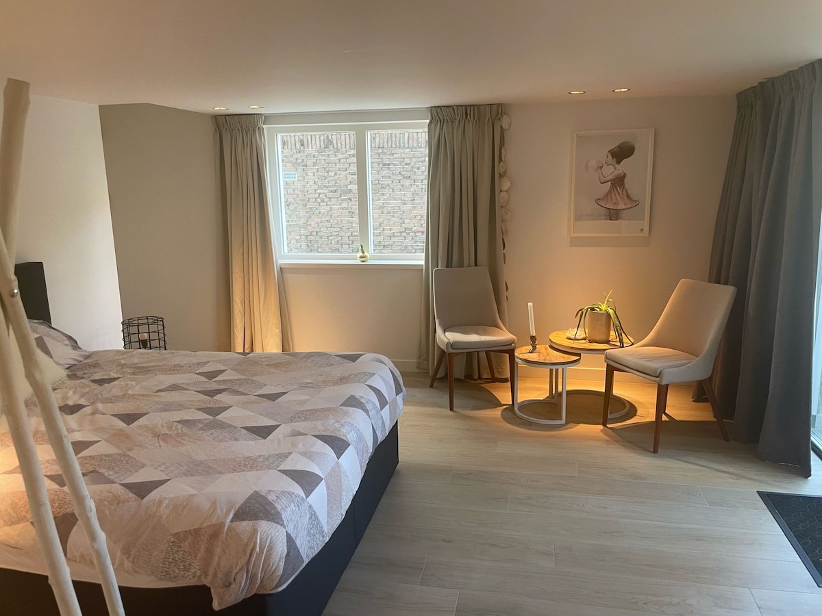 A spacious bedroom is furnished with a large bed, accompanied by two comfortable chairs positioned near a small table. Natural light enters through a large window, revealing soft curtains that frame the view. A piece of artwork adorns the wall beside the seating area.