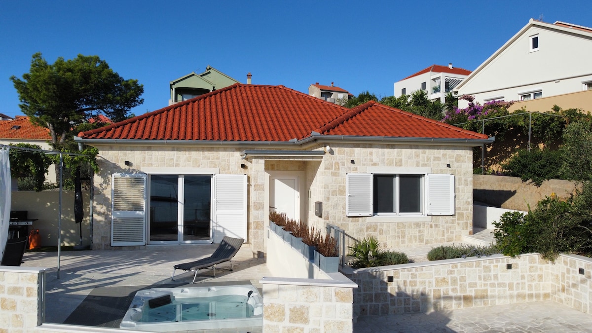 The villa is set against a clear blue sky, featuring a light-colored stone exterior and a red-tiled roof. A spacious terrace includes a whirlpool and a sun lounger, while large glass doors open to the interior, creating a seamless flow between indoor and outdoor spaces.