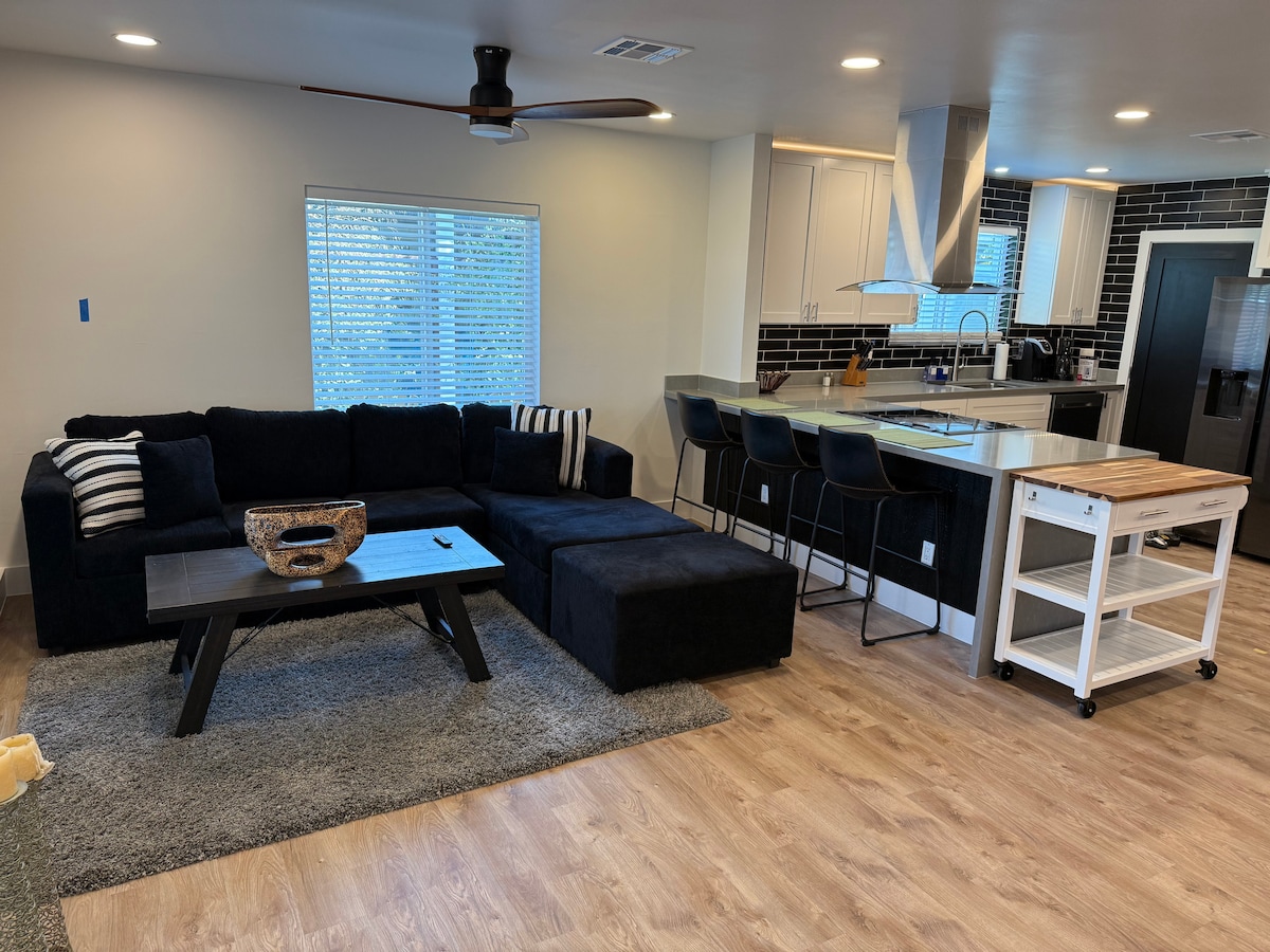 A modern living area features a dark sectional sofa with accent pillows and a low coffee table in front, situated on a soft area rug. In the background, a sleek kitchen showcases a large island with bar stools, complemented by stylish backsplash tiles and ample natural light from a nearby window.