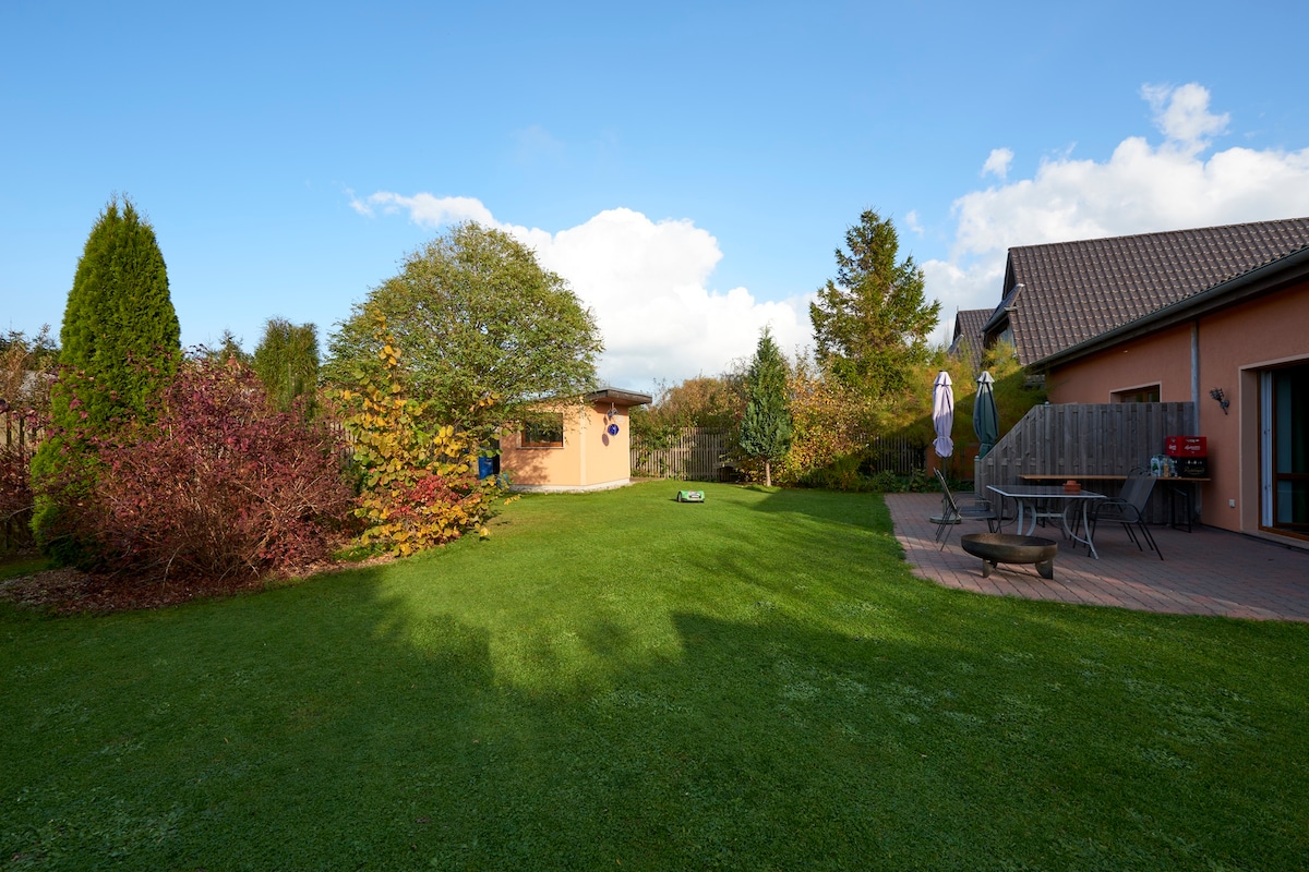 A spacious garden area is bordered by vibrant greenery and seasonal plants. A patio offers outdoor seating with a table and chairs, while a small storage shed is visible in the background. Sunlight creates soft shadows across the lush grass.