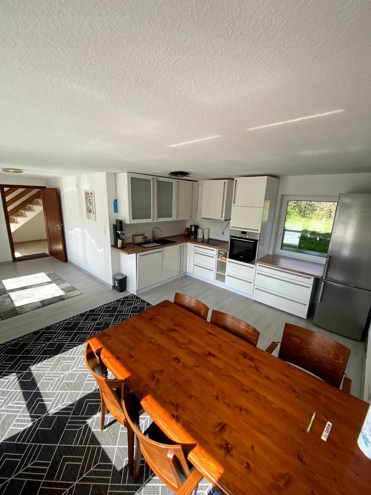 A modern kitchen is featured, equipped with white cabinetry and a wooden counter. A large dining table is set with several chairs. Natural light enters through a window, illuminating the space and highlighting the clean lines and functional design.