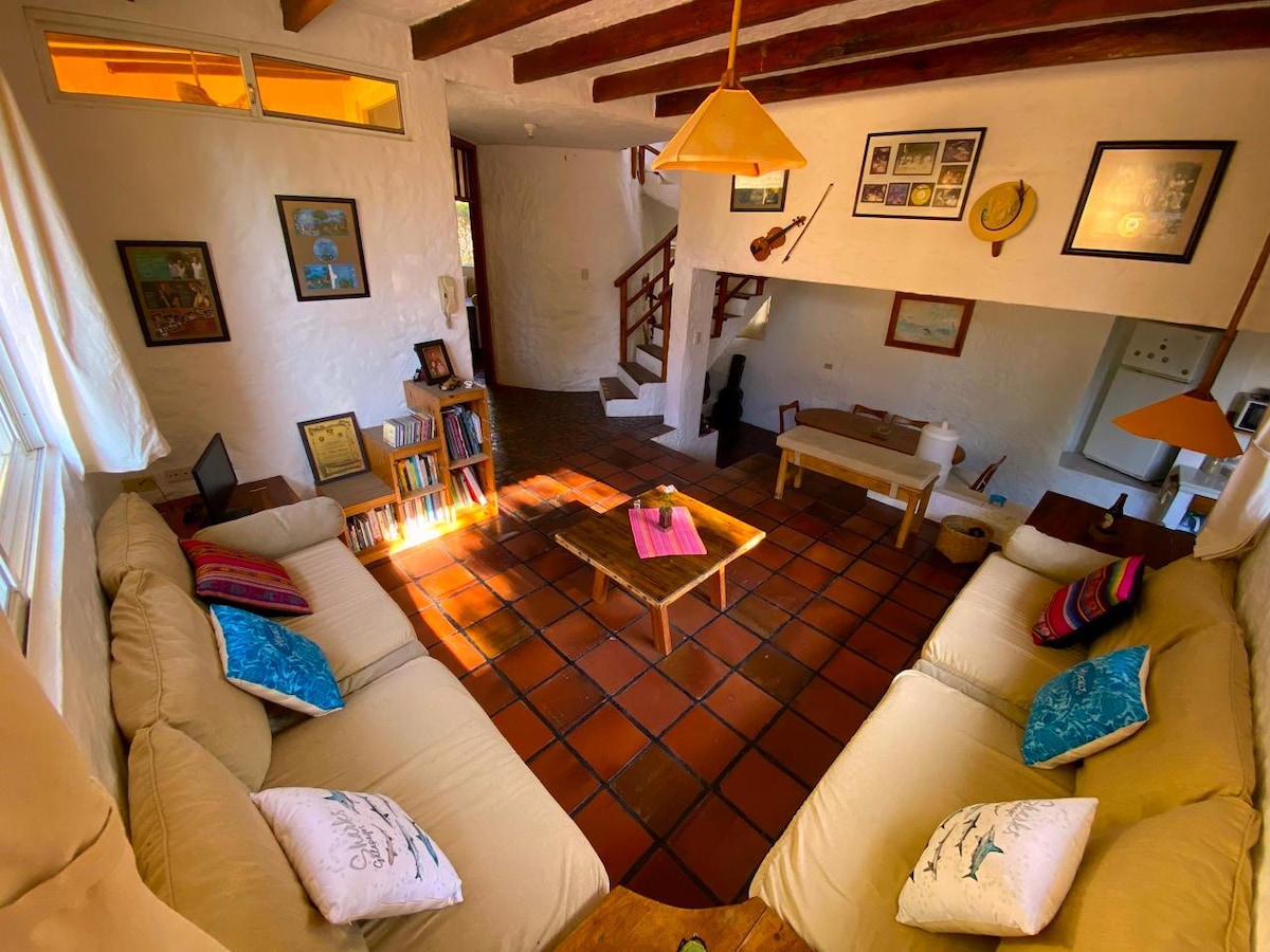 A comfortable living area features a large sectional sofa adorned with colorful cushions. A wooden coffee table is centrally placed, and framed artwork is displayed on the walls. Warm light filters through the windows, creating an inviting ambiance.