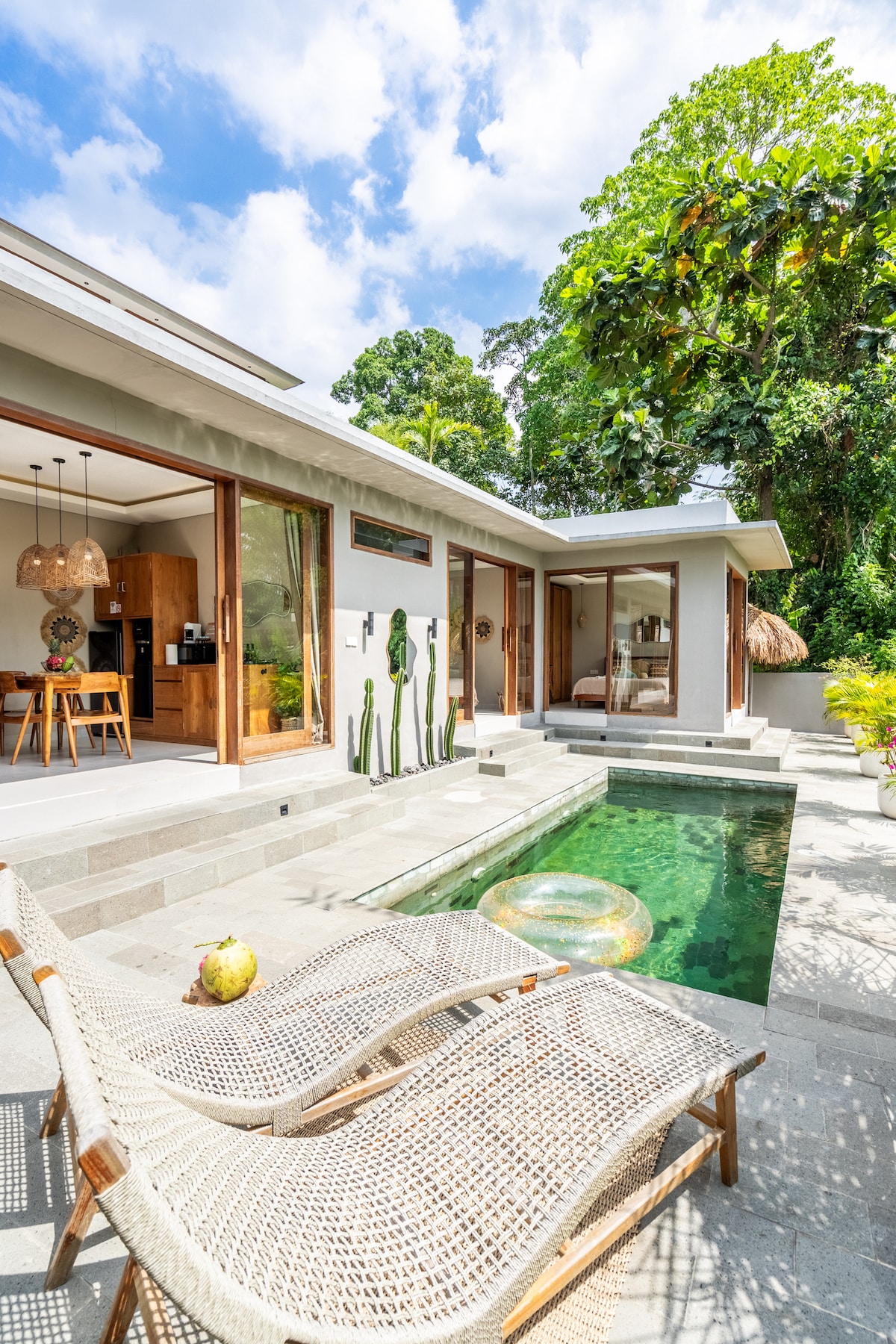 A serene poolside view features two woven lounge chairs positioned beside a clear green pool. Lush greenery and a large tree provide a natural backdrop, while the villa's modern architecture is enhanced by large glass doors and a stylish wooden kitchen visible through the open space.