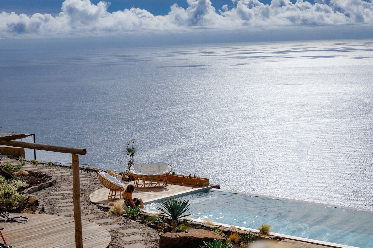 An inviting infinity pool stretches along the cliffside, reflecting the blue ocean and sky. Two comfortable chairs are positioned nearby, surrounded by a natural landscape characterized by various plants and stone pathways. Soft sunlight illuminates the scene, enhancing the serene atmosphere.