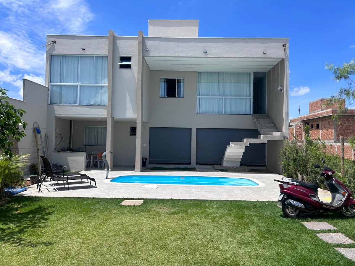 The exterior of the home features a sleek design with large glass windows allowing ample natural light. A private pool glimmers in a well-kept garden surrounded by lush grass. A shaded area with lounge chairs invites relaxation, while the nearby staircase leads to the upper level.