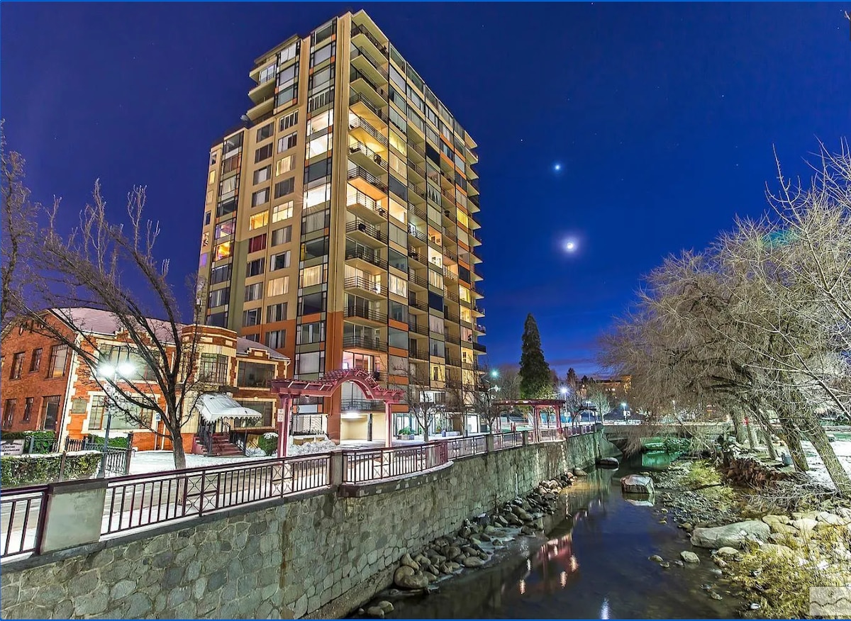 A modern high-rise building stands illuminated at night alongside a flowing creek. The structure features multiple floors of large windows, reflecting the night sky. Trees line the pathway, creating a serene atmosphere in the surrounding area.
