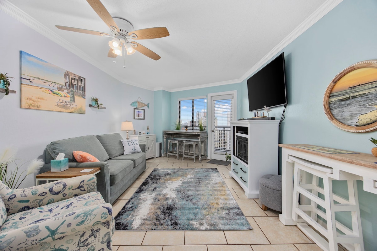The living area features light blue walls with coastal decor and a ceiling fan. A comfortable gray sofa and a patterned area rug are positioned on a tile floor. Natural light enters through multiple windows. A flat-screen TV is mounted on the wall, and a small dining table is seen in the background.