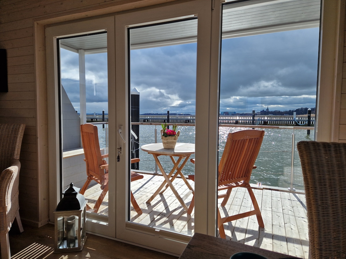 A spacious terrace is shown through large glass doors, featuring a small round table surrounded by two wooden chairs. A vase of flowers sits on the table, while water views and distant buildings add to the serene atmosphere.