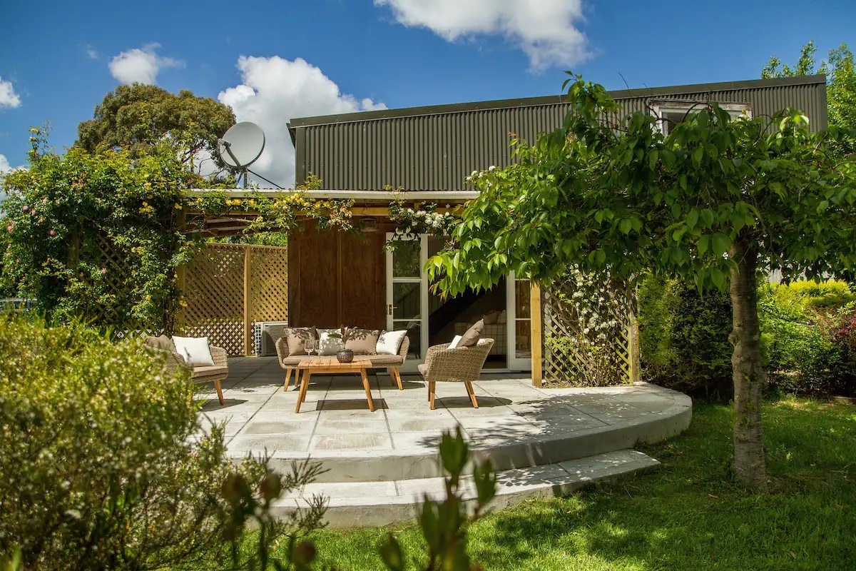 An outdoor seating area features a circular concrete patio adorned with wooden furniture. Lush greenery surrounds the space, with climbing plants crawling on trellises. A two-story building with large windows stands in the background under a partly cloudy sky.