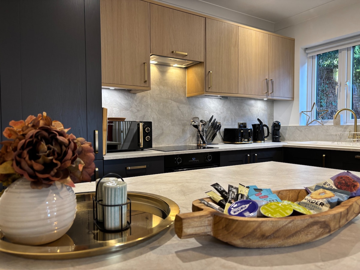 A modern kitchen with light wood cabinetry and a marble countertop showcases sleek appliances, including a microwave and coffee machine. A decorative wooden tray holds an assortment of snacks, while a small vase with a flower adds a touch of warmth. Natural light filters through the window.