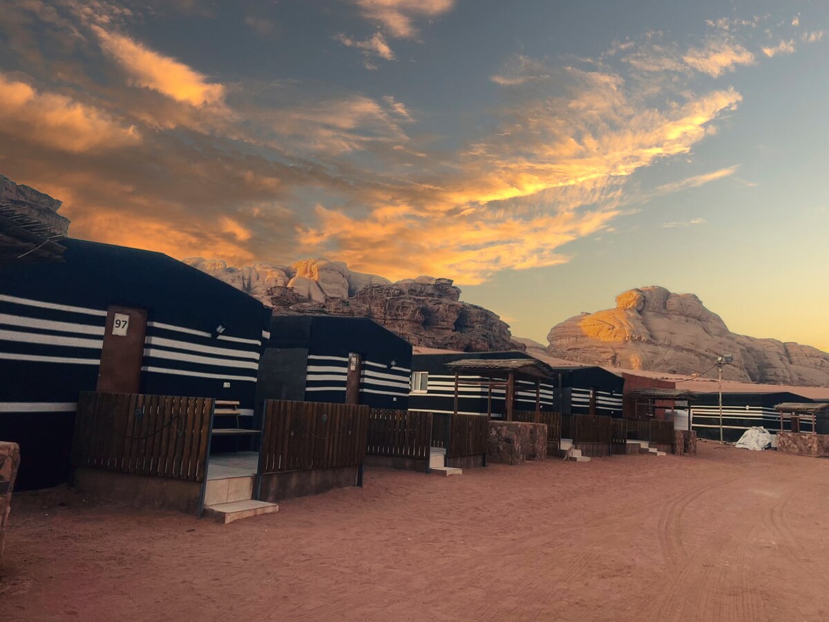 A cluster of traditional Bedouin tents is set against a backdrop of majestic rock formations and a colorful sunset. The tents feature dark exteriors with white stripes, and the sandy ground is visible, adding to the desert ambiance.
