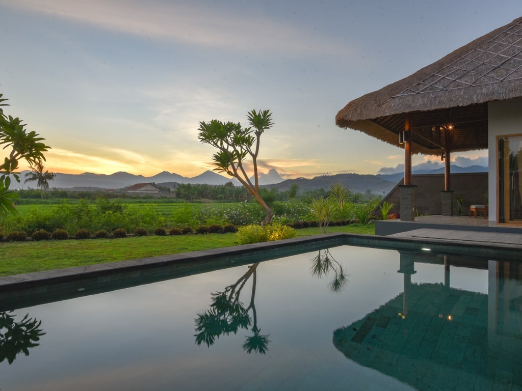 A tranquil private pool reflects the surrounding landscape, framed by lush greenery and distant hills. A traditional thatched-roof structure is positioned nearby, offering a relaxed aesthetic in the peaceful setting. The sky is adorned with soft hues, indicating a serene sunset.