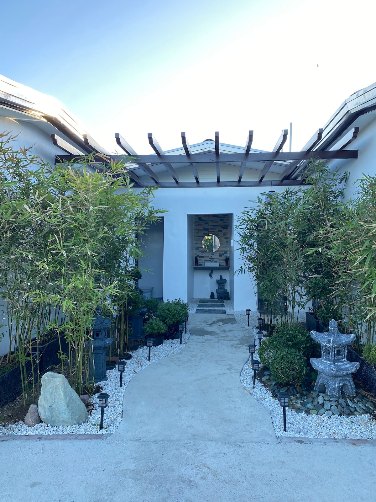 A serene entrance pathway is framed by bamboo and carefully arranged greenery. White gravel lines the path, leading to a subtle sculpture visible in the distance, complemented by stone lanterns along the sides. A soft, natural light illuminates the welcoming approach.