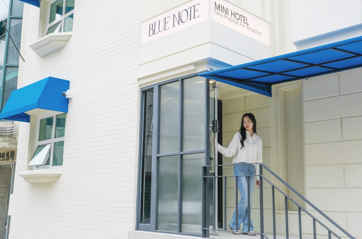 The exterior of Blue Note mini hotel is presented, featuring a stylish entrance with a blue awning. The entrance is framed by large glass windows, allowing ample natural light. A figure stands on the staircase, providing a welcoming presence to the hotel.