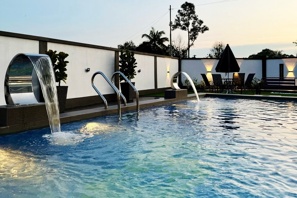 A private pool features smooth water flows from two metallic fountains, creating a calming effect. Surrounding spaces include shaded seating under an umbrella and lounge chairs for relaxation. Lush greenery is visible along the perimeter, complementing the serene atmosphere.