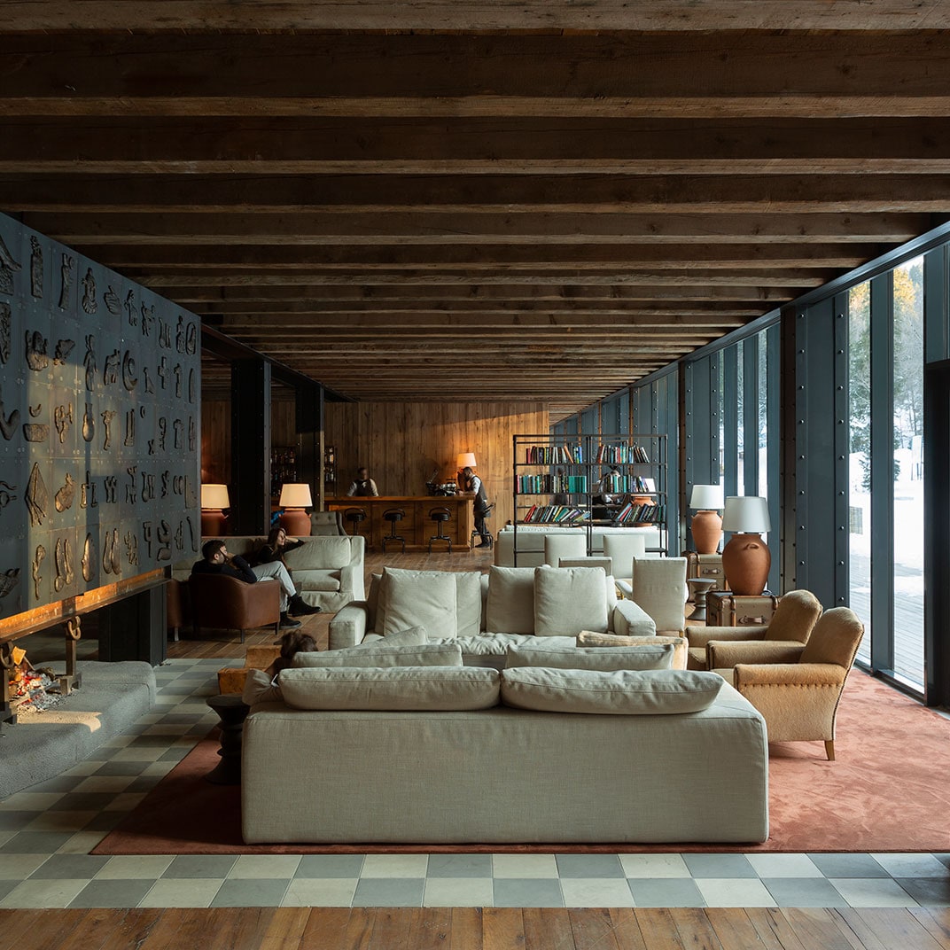 A spacious living area is highlighted by wooden beams and large glass windows that invite natural light. Comfortable sofas are arranged around a central seating area, featuring a warm rug and side tables. A bookshelf is visible, adding a cozy touch to the elegant design.