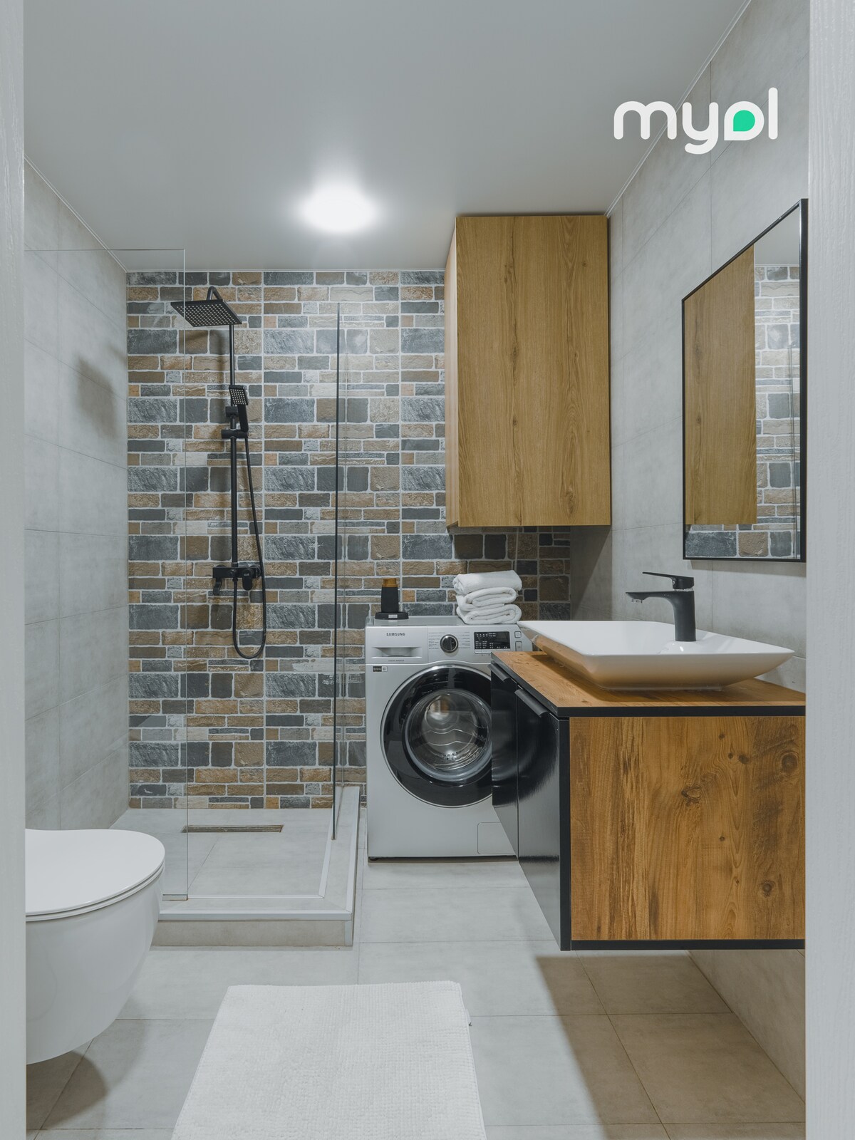 The bathroom features a modern design with a walk-in shower surrounded by tiled walls in earthy tones. A washing machine is positioned beside a wooden cabinet, and a sleek sink is mounted above a storage unit. A white rug is placed on the tiled floor.