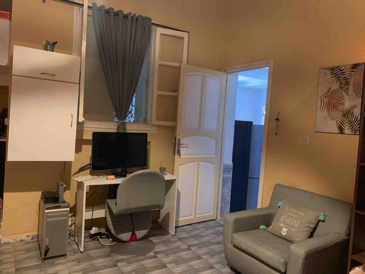 A compact workspace is seen featuring a desk and a monitor. A comfortable chair is positioned nearby. A door opens to another room, while a small refrigerator is visible in the background. Soft lighting enhances the warm yellow walls.