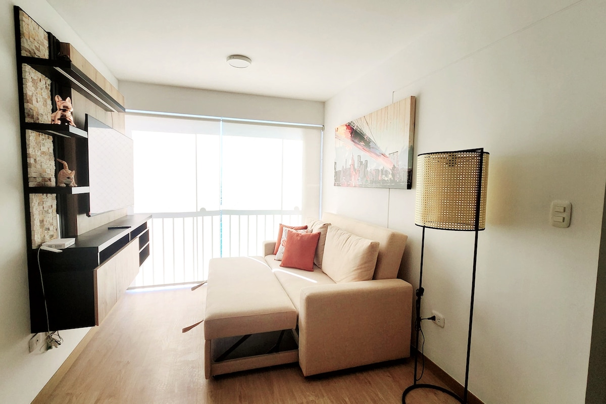 A modern living area features a light-colored sofa bed and a decorative pillow, positioned near a large window. A floor lamp stands beside the sofa, providing additional lighting. Wall-mounted shelves display art and decorative items, contributing to a contemporary design.