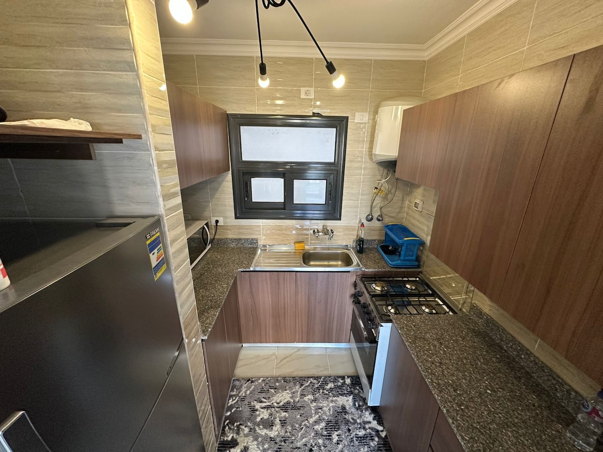 A modern kitchen space features granite countertops and wooden cabinetry. Stainless steel appliances, including a refrigerator and a gas stove, are present. A window allows natural light, enhancing the functional layout. A patterned rug lies on the floor, adding subtle texture to the environment.