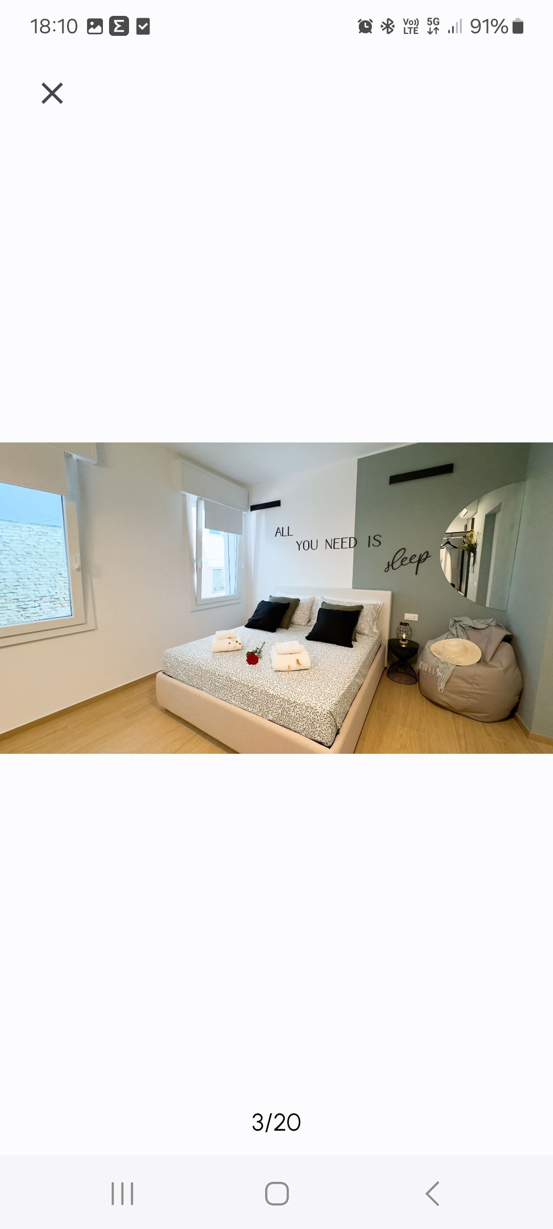 A well-appointed sleeping area features a spacious bed made with light-coloured linens. Black accent pillows add contrast. Natural light filters through two windows with minimalist treatments. A cozy bean bag chair is positioned nearby, complementing the modern design.