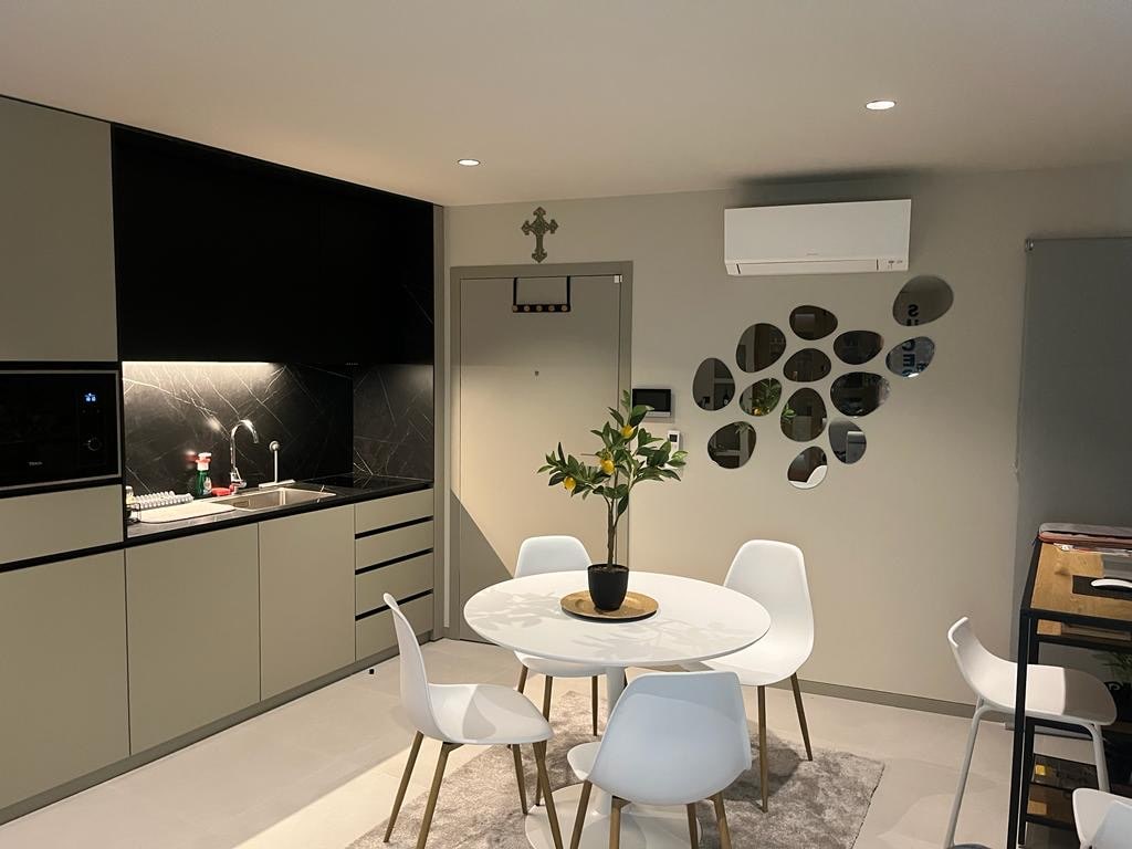 A modern kitchen area is featured, showcasing sleek cabinetry and a black marble backsplash. A round dining table is surrounded by white chairs, and a potted plant adds a touch of nature. Soft illumination enhances the contemporary design, with a mirror decor enhancing the space.