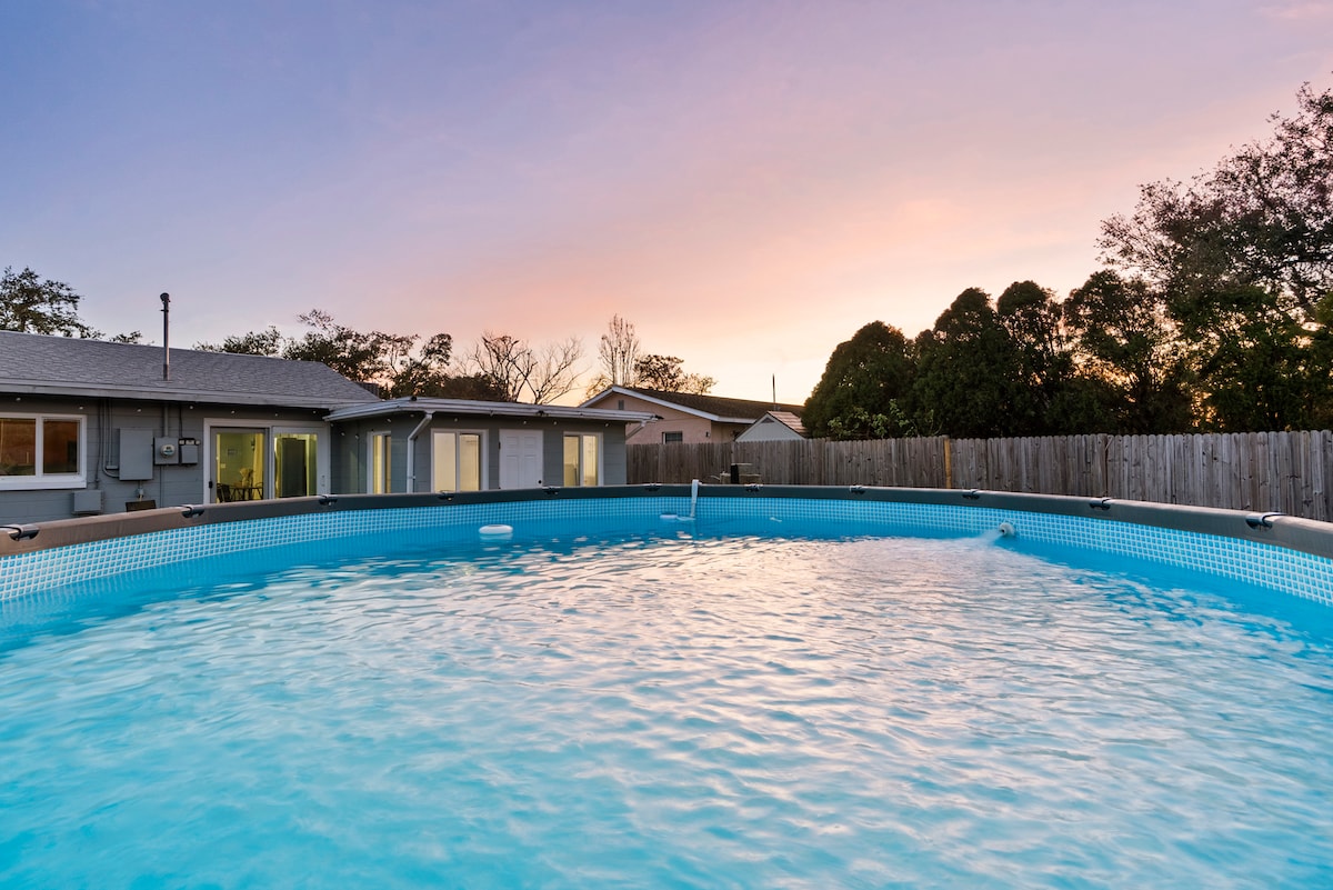 An above-ground pool is visible, set against a backdrop of a colorful sunset sky. The surrounding area features a privacy fence and trees, creating an inviting outdoor space for relaxation.