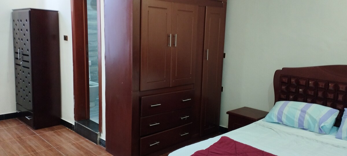 A comfortable sleeping area features a spacious bed with a simple, quilted headboard and decorative pillows. A wooden wardrobe and a bedside table are positioned nearby. A bathroom doorway is visible in the background, indicating easy access to additional amenities.