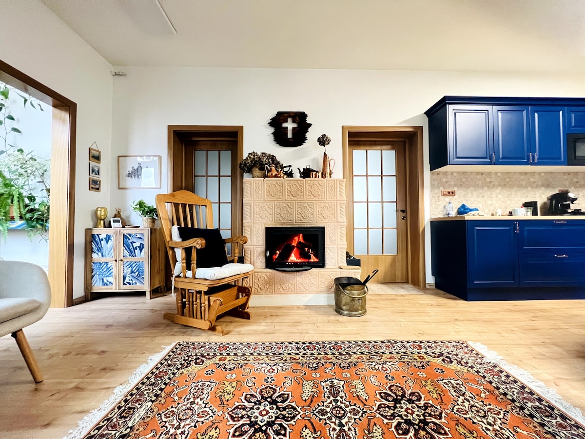A cozy living area features a wooden rocking chair positioned near a small, brick fireplace with a lit fire. The kitchen area can be seen in the background, equipped with deep blue cabinetry and a range of kitchen appliances. A decorative area rug adds warmth to the space.