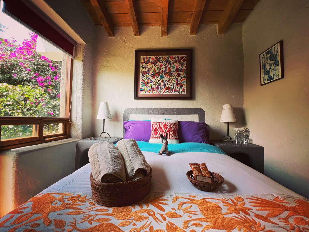 A cozy sleeping area features a spacious bed adorned with colorful pillows and a patterned blanket. Natural light filters through the window, highlighting a basket with towels and snacks arranged on the bed. Vibrant flowers can be seen outside, enhancing the room's inviting ambiance.