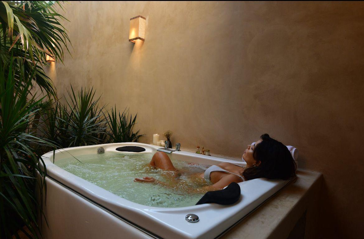 A private hot tub is surrounded by lush greenery and softly lit wall sconces, creating a serene atmosphere. The water appears inviting, reflecting soft light. Various plants add to the relaxed environment, while the surrounding walls feature a warm earthy tone.