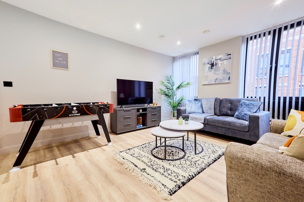 The living area features a foosball table, a large television on a dark entertainment unit, and two comfortable sofas. A stylish coffee table rests on a patterned rug, with natural light filtering through vertical blinds on the windows. Decorative plants add a touch of greenery.