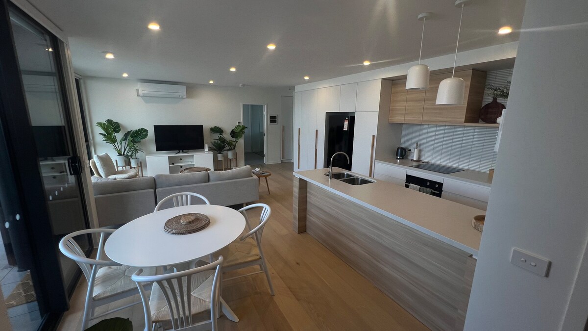 An open plan living area features a kitchen with modern appliances and a sleek countertop. A dining table with chairs is positioned nearby, while a comfortable seating arrangement is visible across the space. Light from the windows enhances the airy ambiance, complemented by indoor plants.