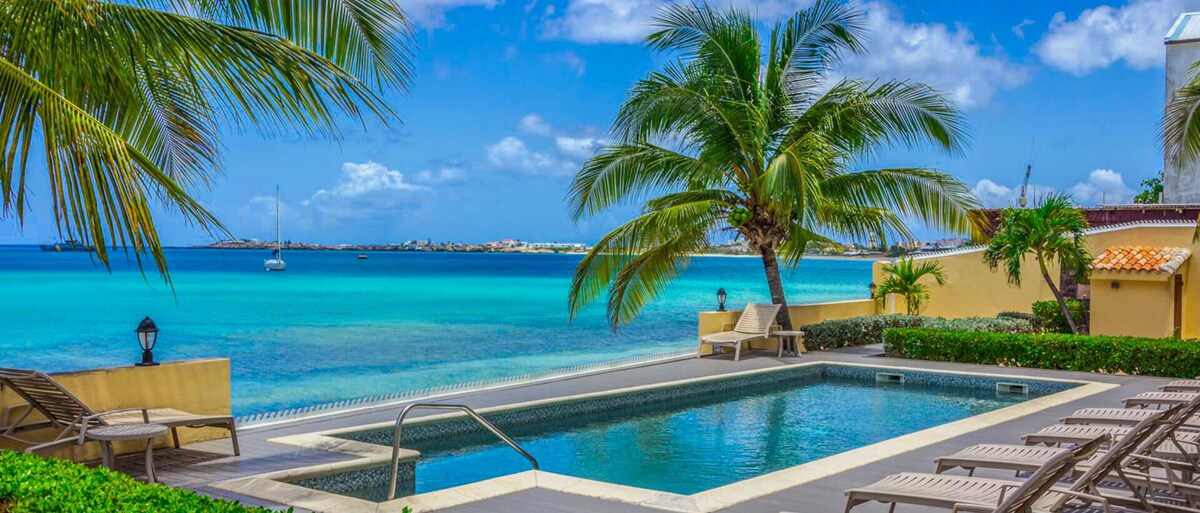 A beachfront pool area is surrounded by lush greenery, featuring several lounge chairs arranged neatly. Tall palm trees sway gently in the breeze, framing a stunning view of clear turquoise waters and a sky dotted with fluffy clouds.