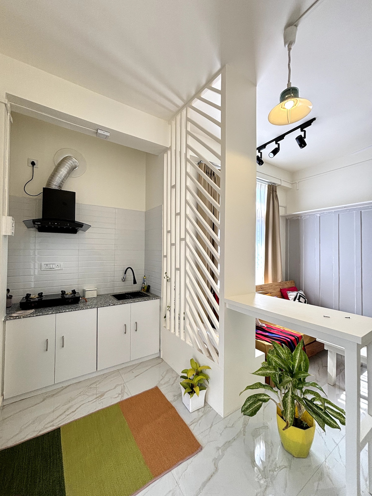 The compact kitchen area features a sleek countertop with a sink and a stovetop, flanked by white cabinets. A small potted plant adds a touch of greenery. Natural light floods from nearby windows into the open living space, enhancing the modern decor.