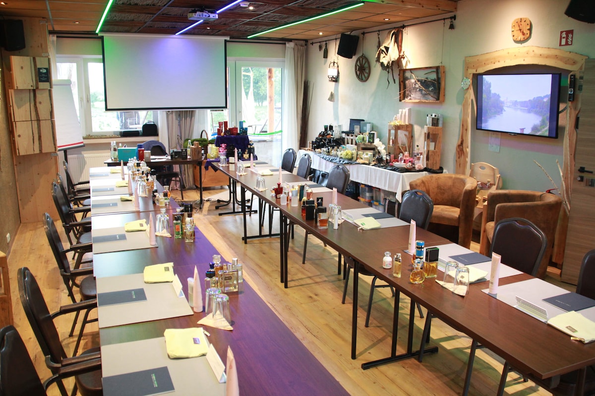A spacious meeting room is set up with multiple tables arranged in a U-shape. Each table is equipped with notepads and pens, along with refreshments. Large screens are positioned for presentations, and soft seating is visible in the background, creating a comfortable environment.