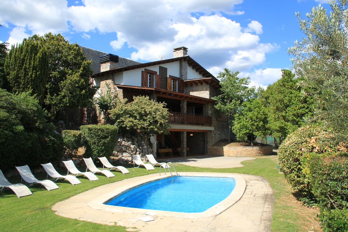 A spacious outdoor area is highlighted by a rectangular pool surrounded by a stone patio. Lounge chairs are arranged neatly nearby, and lush greenery frames the house, which showcases a blend of natural stone and wooden accents. Large windows create a welcoming atmosphere.