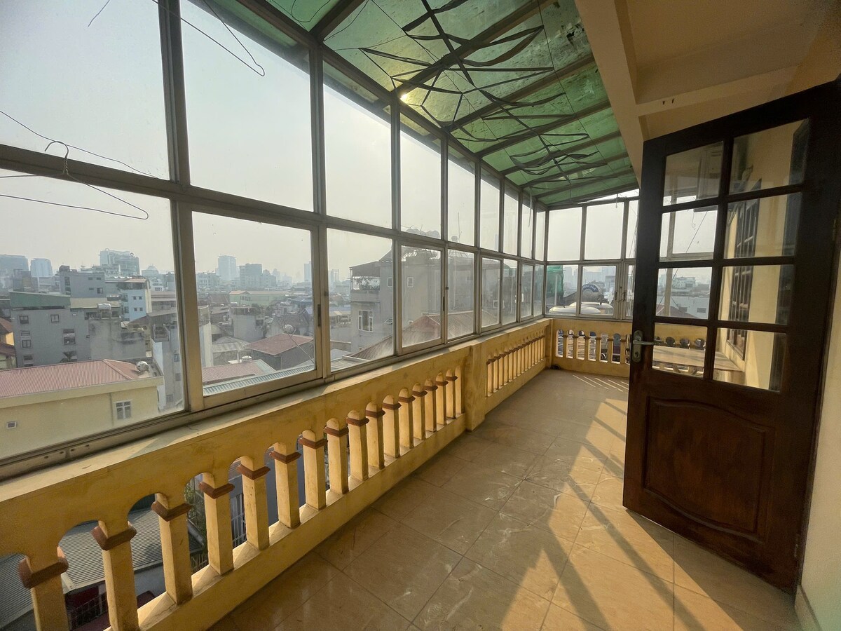 A spacious balcony is filled with natural light, featuring floor-to-ceiling windows that offer unobstructed city views. The tiled floor contrasts with wooden railings, creating a serene space perfect for relaxation. Light and shadow interplay highlight the architectural details throughout the area.