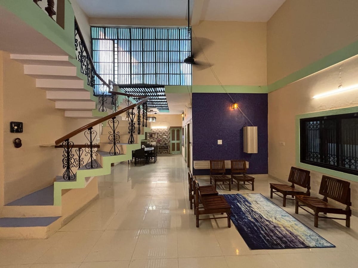 An open common area is presented, featuring a staircase with decorative railing and multiple seating options. Large windows allow natural light to stream in, illuminating tiled floors. A vibrant rug is laid out in the seating area, creating a welcoming space for guests to relax.