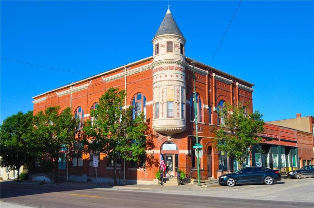A historic brick building is featured, showcasing intricate architectural details, including a turret and arched windows. Lush greenery surrounds the structure, and vehicles are parked along the street. A clear blue sky serves as a backdrop, enhancing the building's vibrant colors.