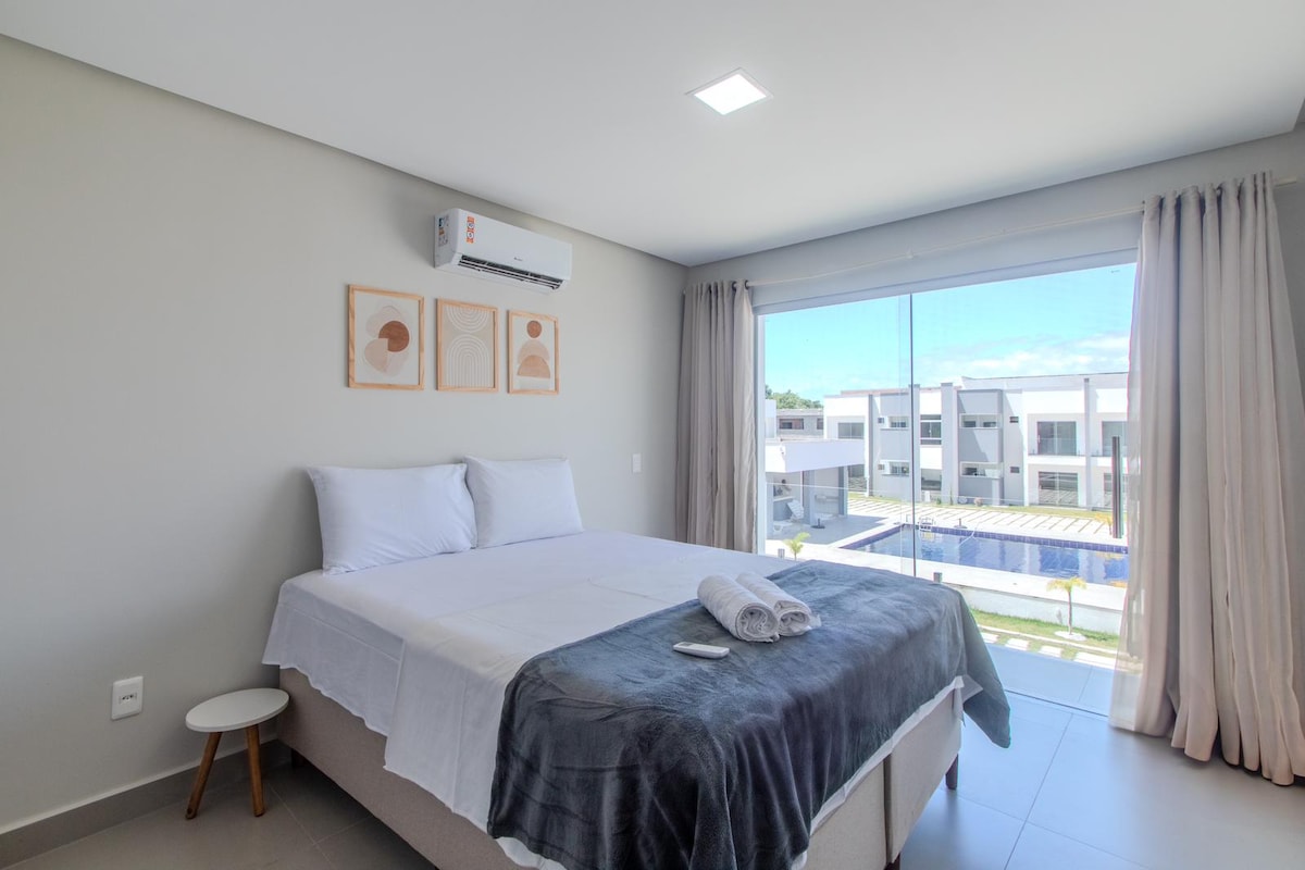A bright bedroom is presented with a spacious bed, complemented by neutral-colored linens and towels. Natural light enters through large windows covered by sheer curtains, offering views of the surrounding area. Air conditioning unit is mounted on the wall for comfort.