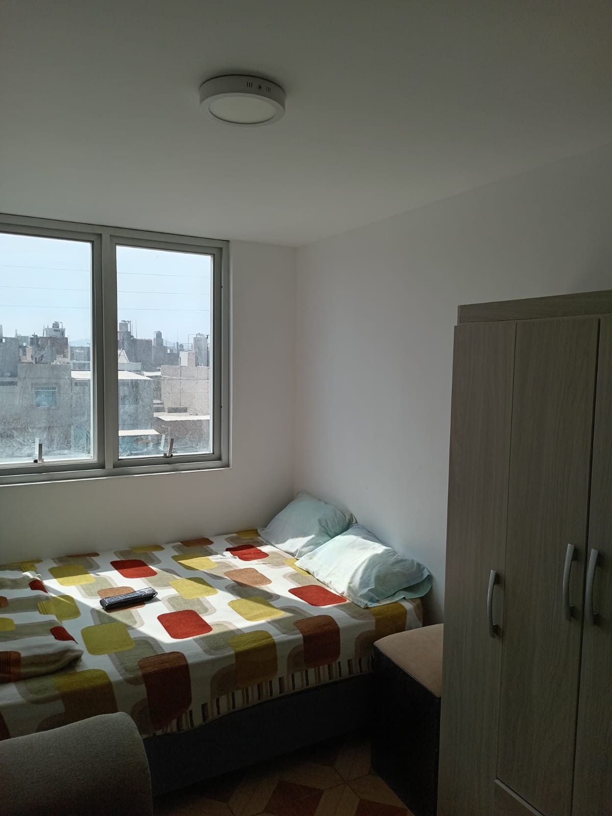 A bright sleeping area features a bed covered with a patterned blanket in warm colors. Two large windows allow ample natural light to fill the room, providing views of the cityscape. A simple wardrobe stands nearby, creating a clean and organized space.