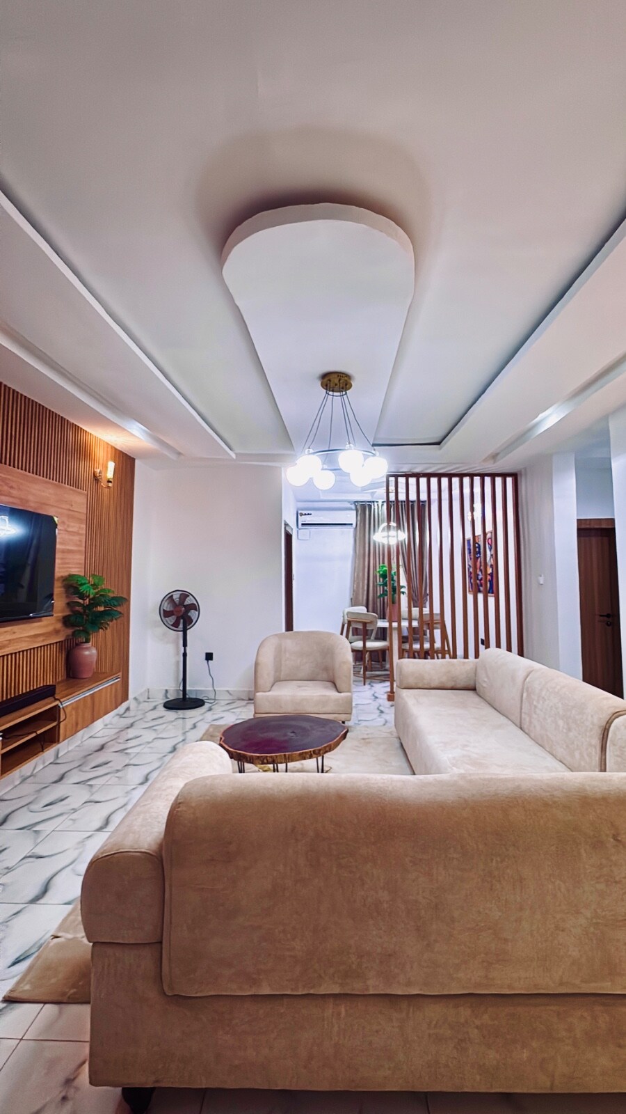 A spacious living area features a soft beige sofa and a central round coffee table. Decorative elements include a tall indoor plant and a stylish ceiling fixture with multiple lights. A wall-mounted television is positioned against a wooden panel, enhancing the room's contemporary design.